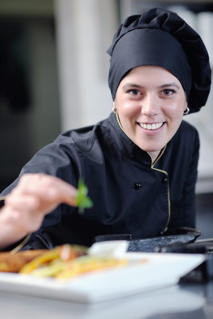 Female chef in black uniform smiling while adding herbs to a freshly plated dish.