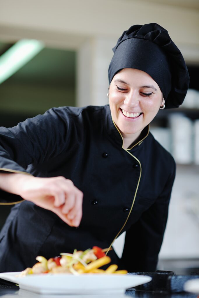 Smiling female chef in black uniform garnishing a colorful pasta dish in the kitchen.